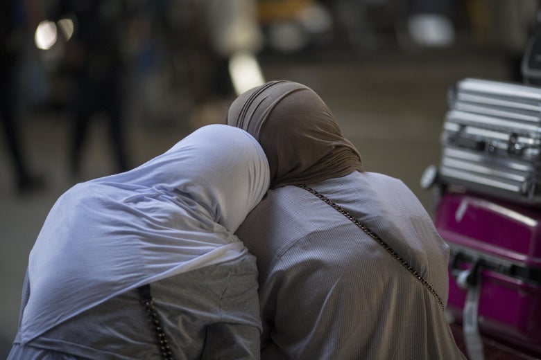 Two Muslim women lean against each other, as seen from behind. Suitcases can be seen next to them.